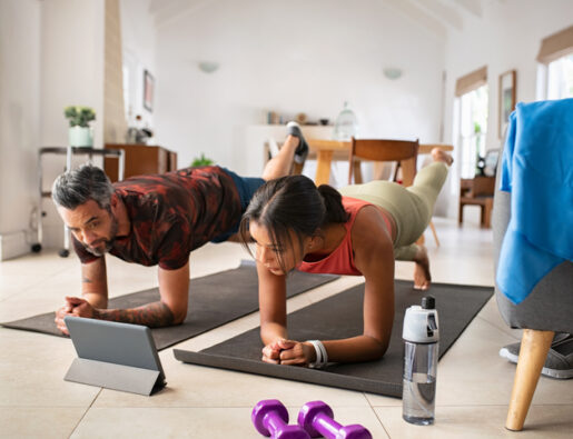 Multiethnic mature couple exercising at home and watching training videos on digital tablet. Middle aged woman and indian man doing planks with a leg outstretched while watching fitness lessons online on digital tablet. Fit mid adult couple doing strenght plank following online tutorials.